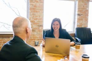 Une femme souriante assise à son bureau en face d'un homme (non visible) souriant également. Elle a un ordinateur portable devant elle en train de réaliser un coaching pour managers.
