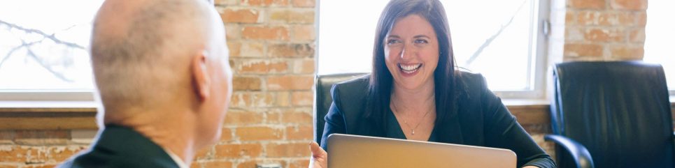 Une femme souriante assise à son bureau en face d'un homme (non visible) souriant également. Elle a un ordinateur portable devant elle en train de réaliser un coaching pour managers.